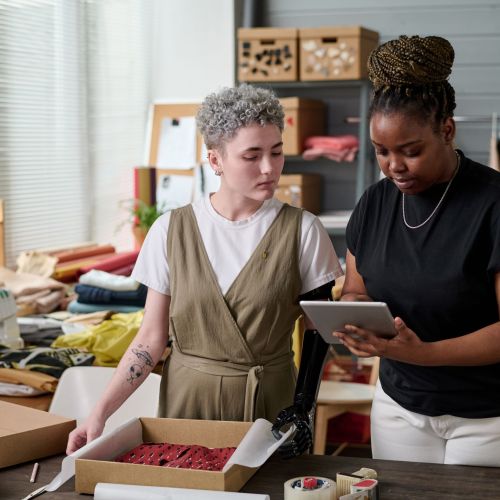 Two young intercultural females checking address of client while packing order, woman with tablet showing data to colleague with disability